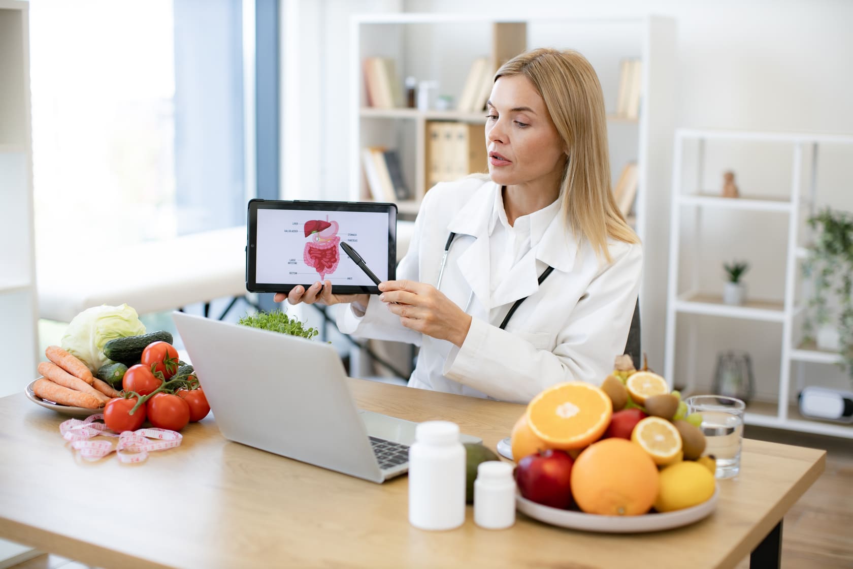 Nutritionist showing digestive system diagram during online consultation. Healthy fruits and vegetables on table.