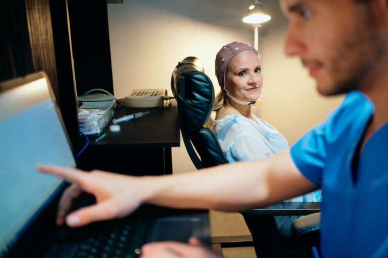 A female executive undergoing a brain function assessment using neuro-monitoring equipment, while a clinician analyzes cognitive data on a screen.