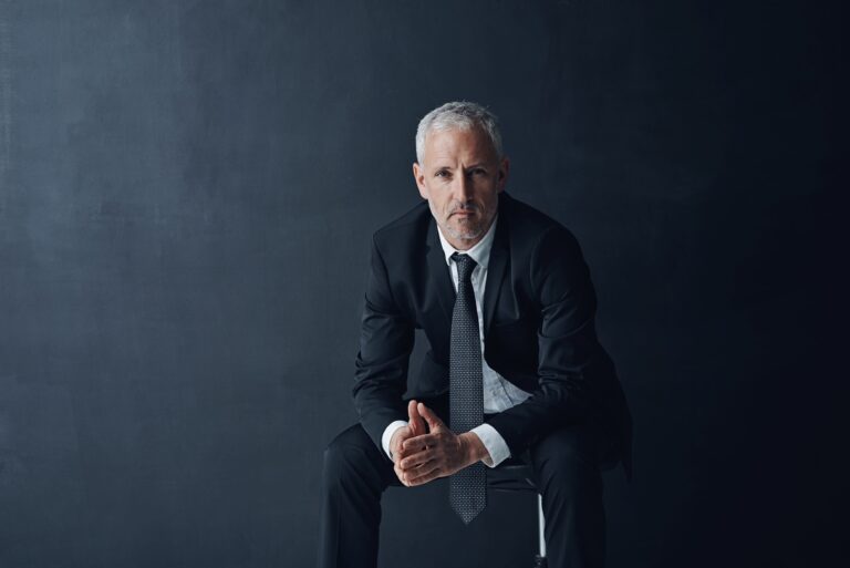 Confident senior male executive in a tailored black suit sitting against a dark backdrop, representing leadership, longevity, and performance-driven health strategies.
